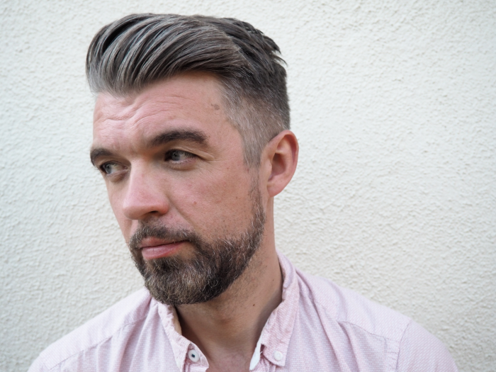 A person in a light pink button-down shirt stands in front of a white wall and looks to the side.